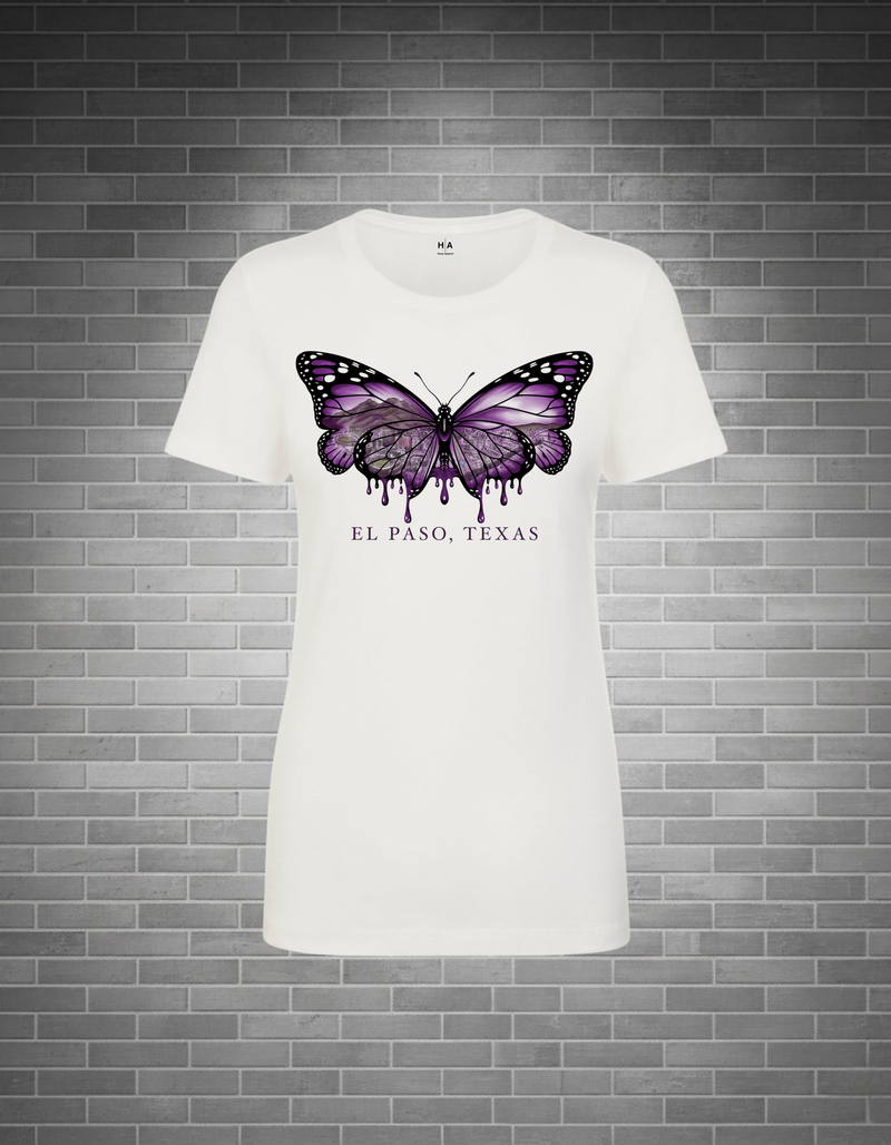 A white short-sleeve crewneck t-shirt featuring a purple and black dripping butterfly graphic on the chest, with 'EL PASO, TEXAS' printed below it. The shirt is displayed against a gray brick wall background.