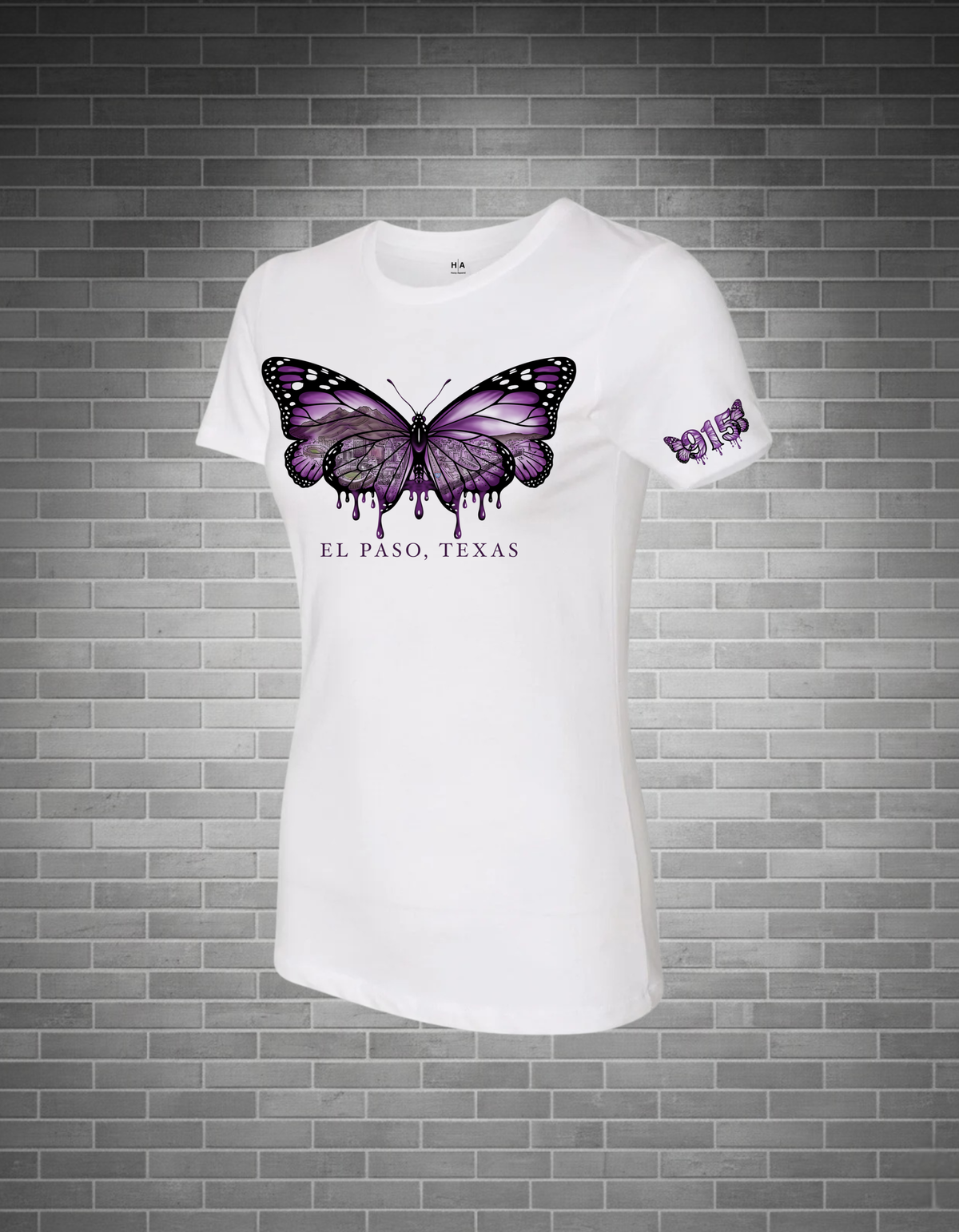 A white short-sleeve crewneck t-shirt featuring a large purple and black butterfly graphic on the chest with 'EL PASO, TEXAS' printed below, displayed against a gray brick wall background.