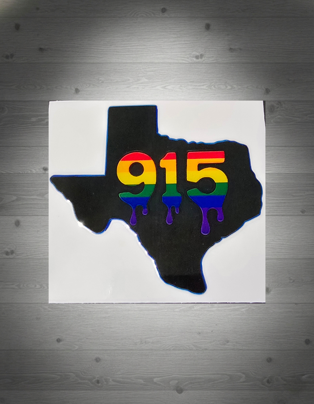 Texas-shaped sticker with rainbow '915' on a grey wooden background
