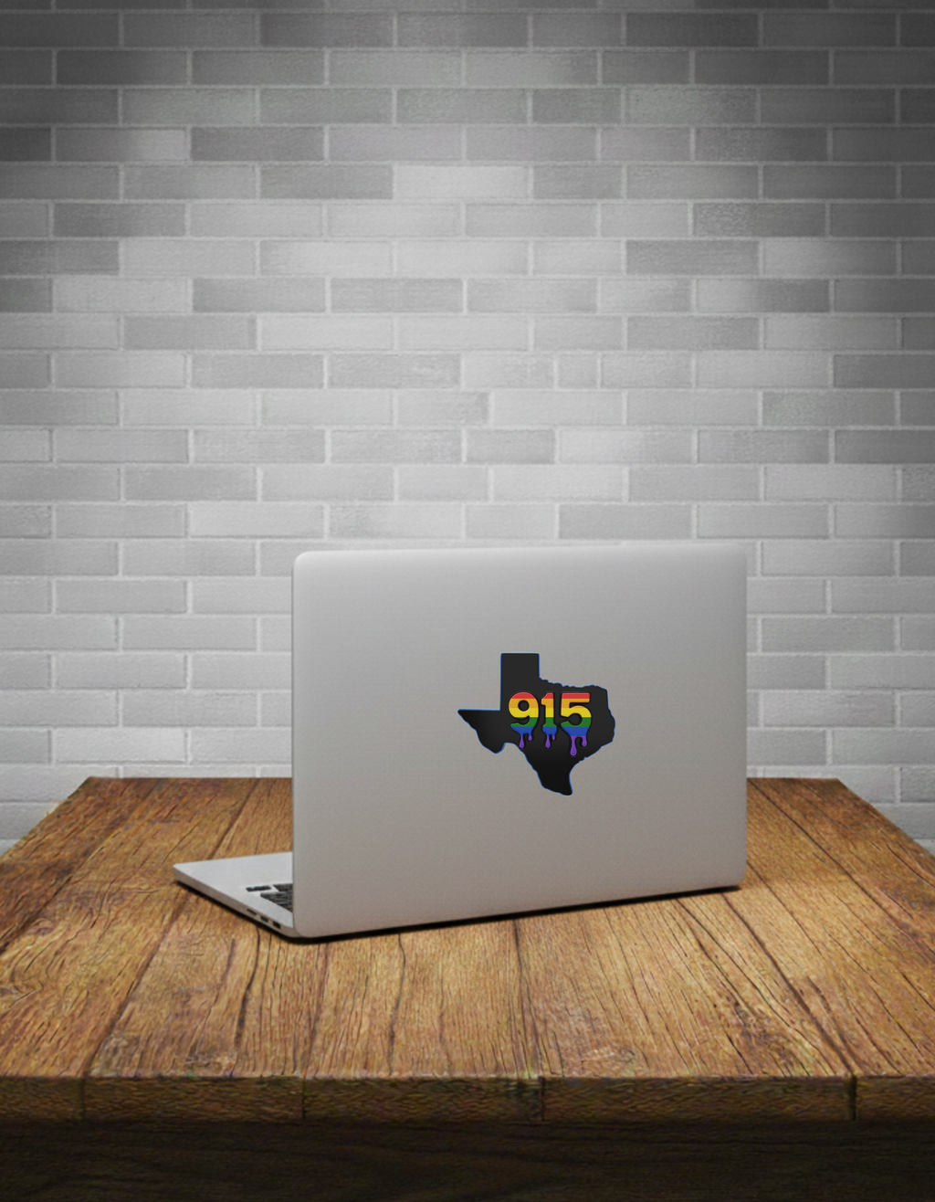 Laptop with a rainbow Texas-shaped sticker on a wooden table against a grey brick wall.