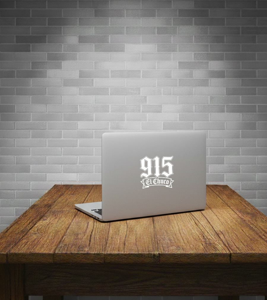 A silver laptop sits open on a rustic wooden table against a gray subway tile wall. The laptop lid has a white graphic decal reading '915 El Chico'.