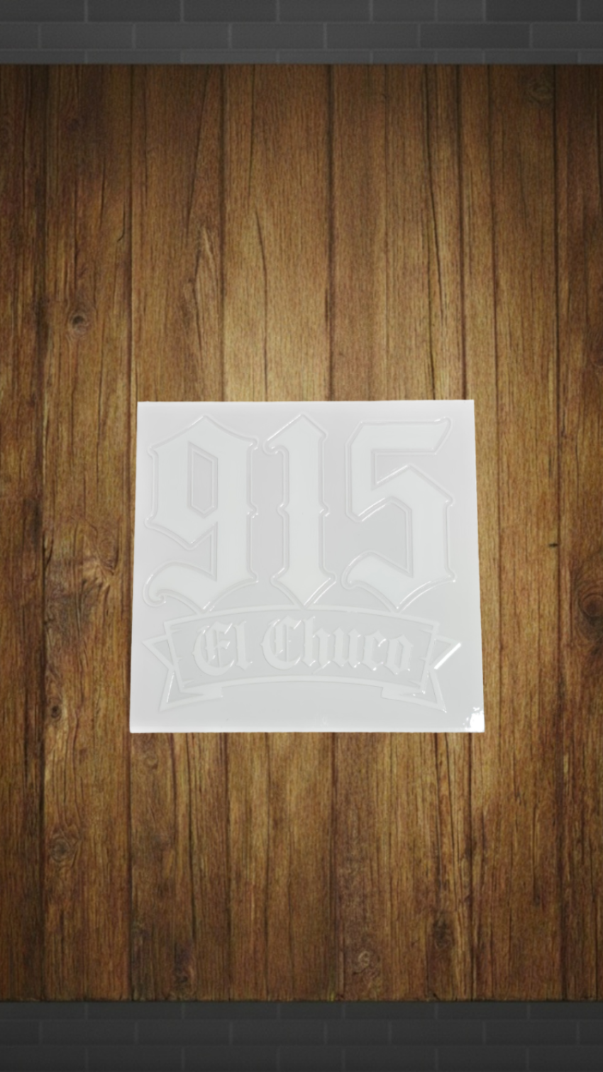 A square white sticker or decal with a faded gray '915 El Clueño' design, shown against a brown wood plank background.