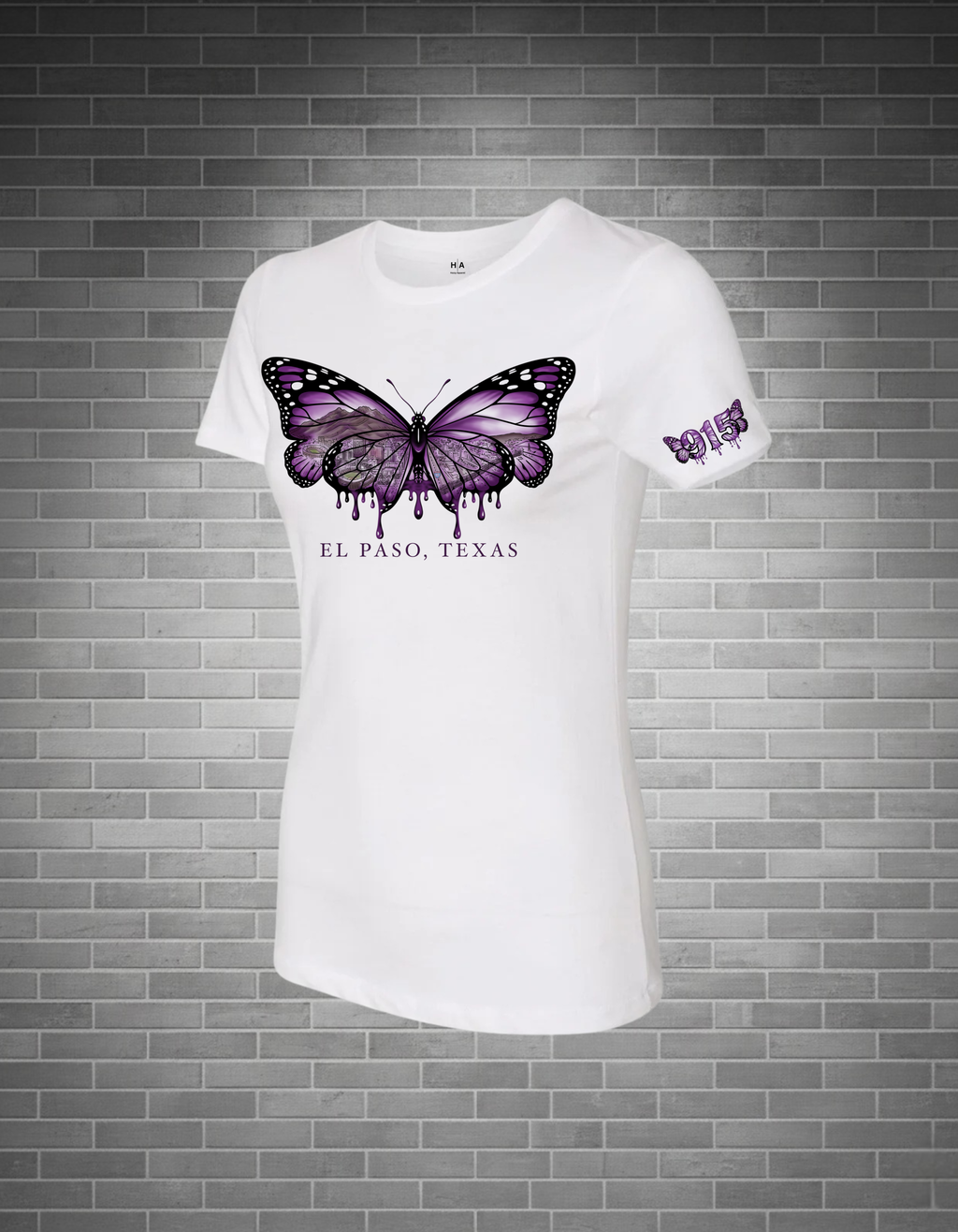A white short-sleeve crewneck t-shirt featuring a large purple and black butterfly graphic on the chest with 'EL PASO, TEXAS' printed below, displayed against a gray brick wall background.
