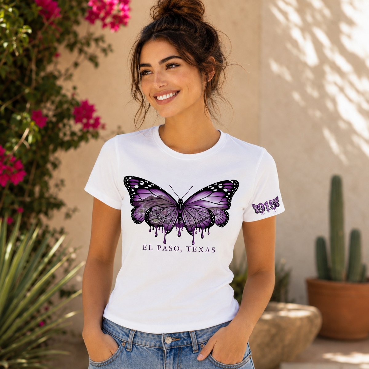 A woman wearing a white crewneck t-shirt with a purple dripping butterfly graphic and 'EL PASO, TEXAS' printed below it, posing outdoors against a beige stucco wall with plants.