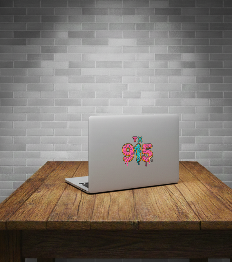 A silver laptop sits open on a rustic wooden table against a gray subway tile wall. The laptop lid features a colorful graphic decal spelling '915' in dripping pink and teal lettering.