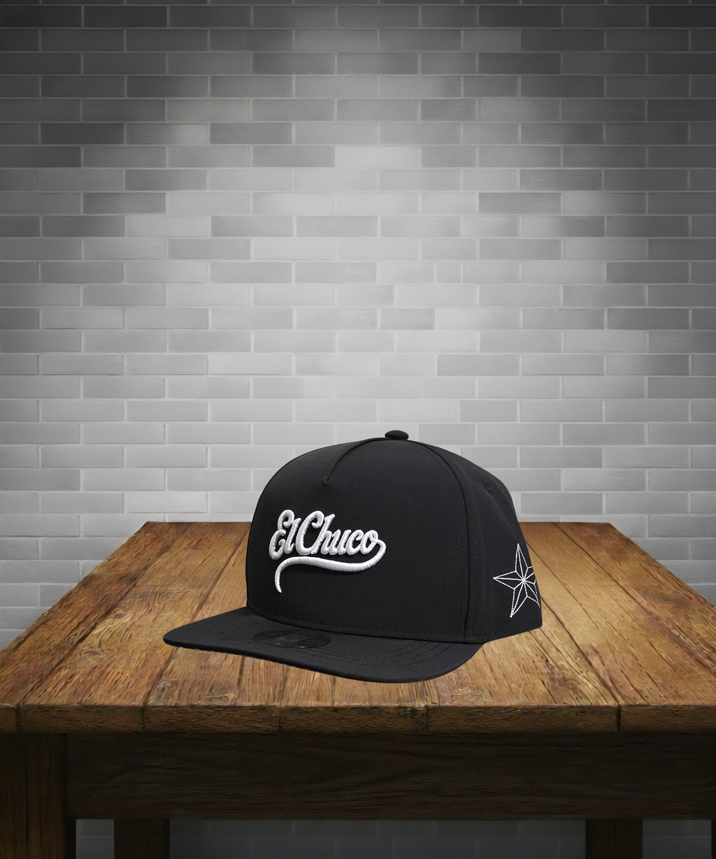 Black cap with 'El Chuco' logo on a wooden table against a gray brick wall.