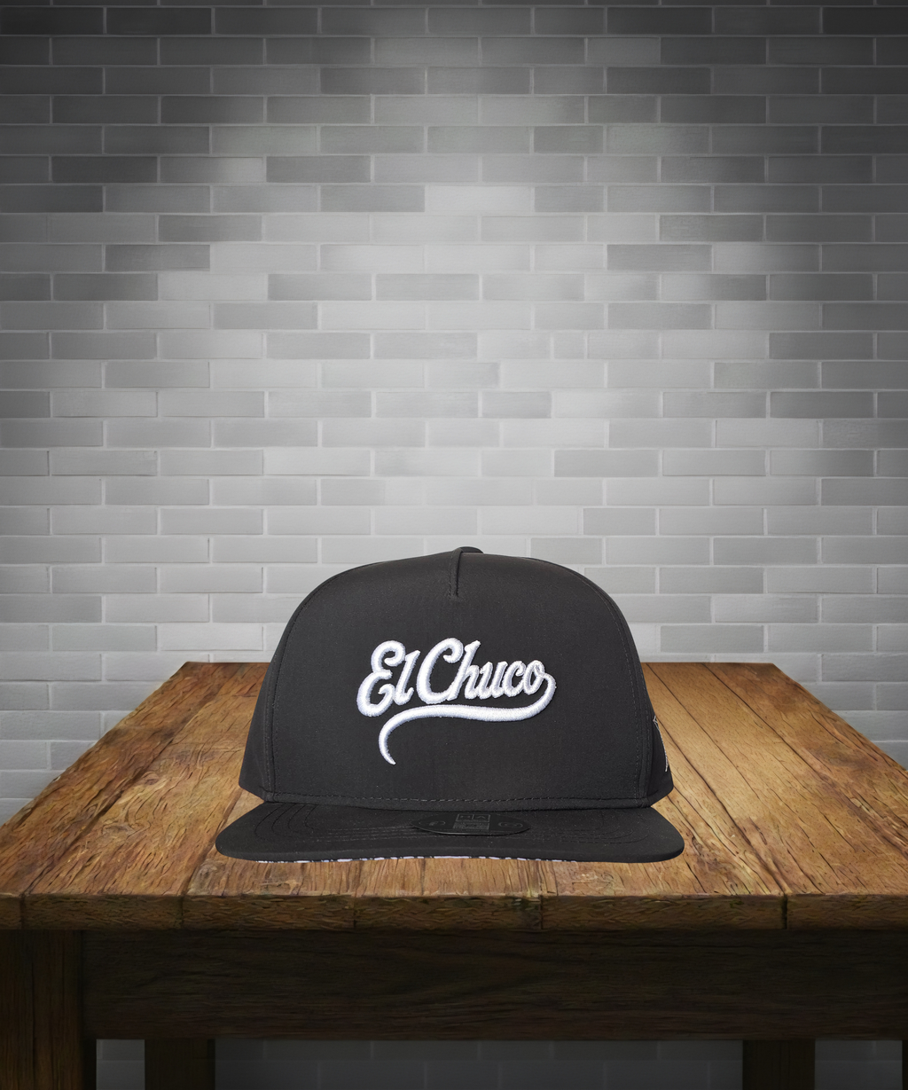 Black cap with 'El Chuco' logo on a wooden table against a gray brick wall.