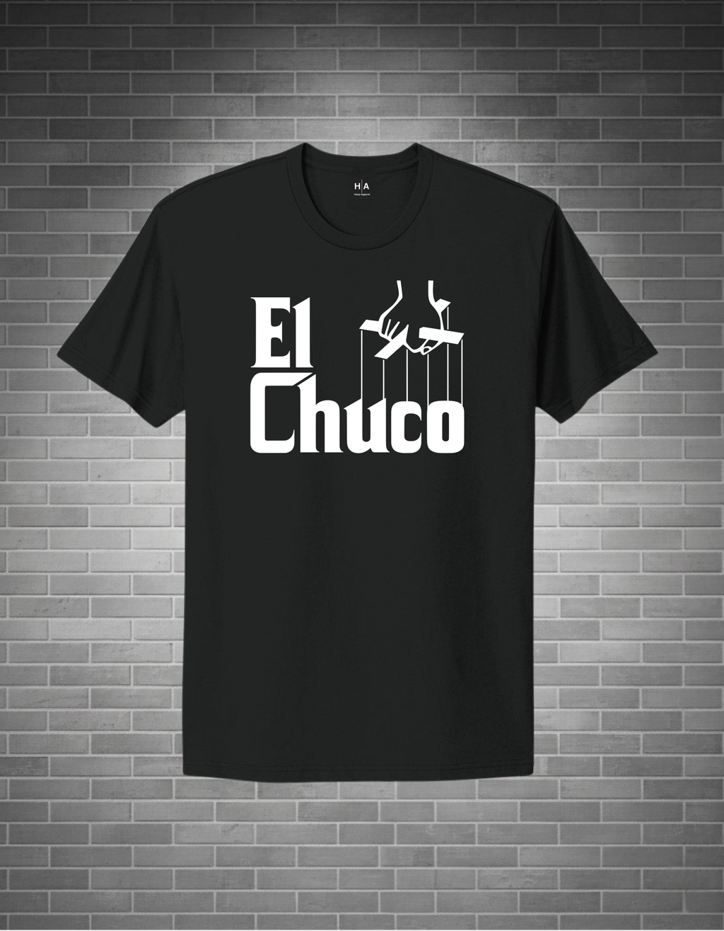 A black short-sleeve t-shirt with a white graphic print reading 'El Chuco' and puppet strings, displayed against a gray brick wall background.