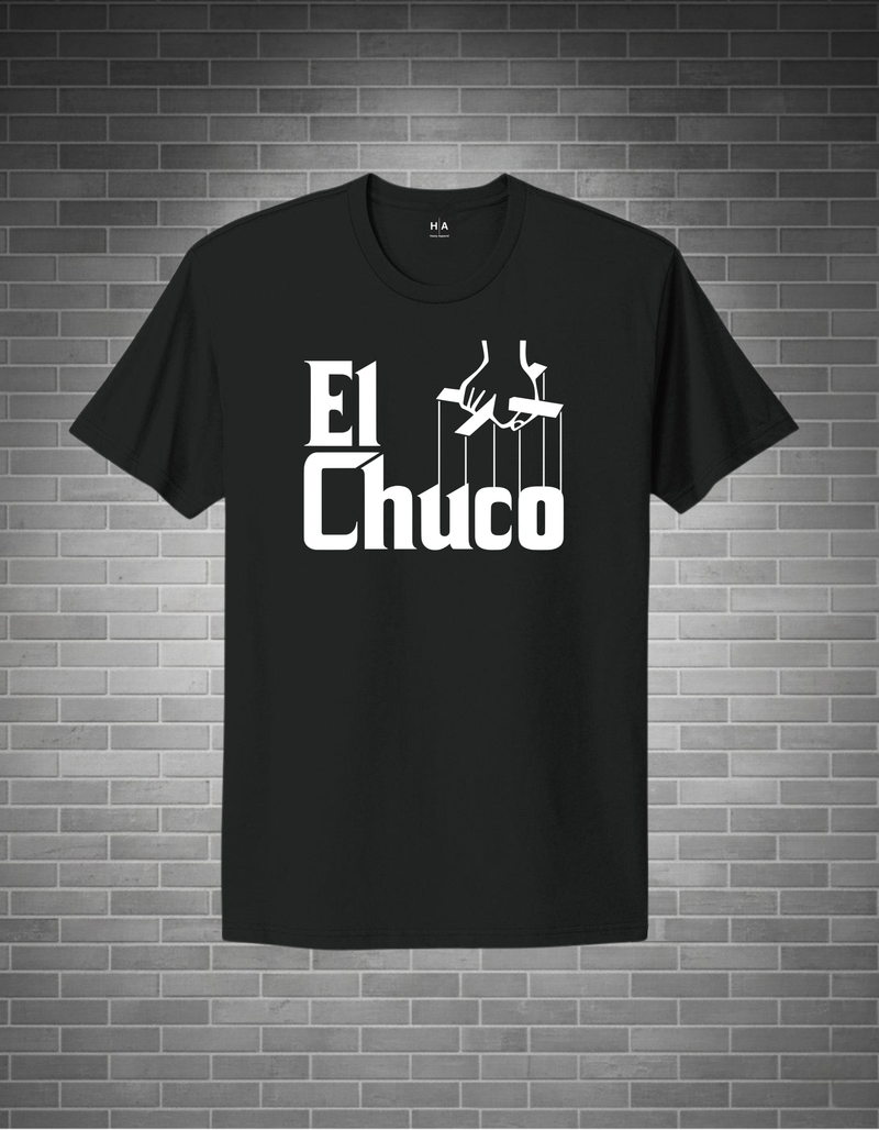A black short-sleeve t-shirt with a white graphic print reading 'El Chuco' and puppet strings, displayed against a gray brick wall background.
