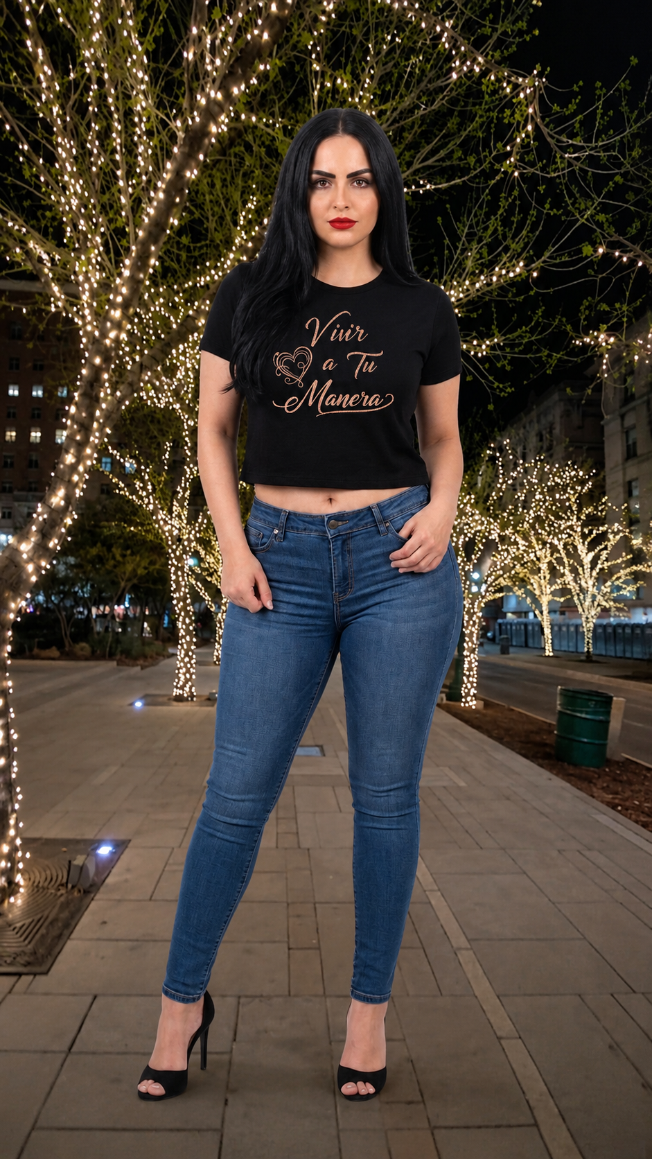 Woman wearing a black t-shirt and blue jeans standing on a sidewalk with decorative lights in the background