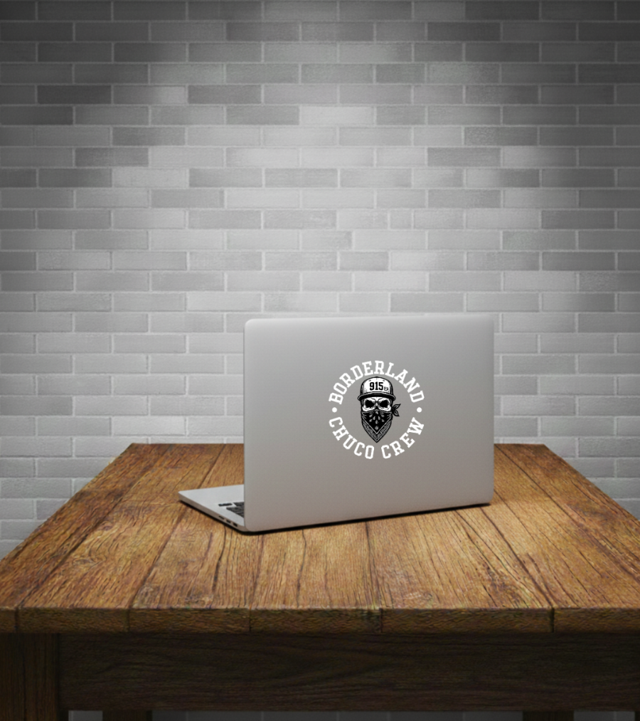 A silver laptop sits open on a rustic wooden table against a gray subway tile wall. The back of the laptop has a circular white sticker reading 'BORDERLAND CHOCO CREW' around a black illustrated face graphic.
