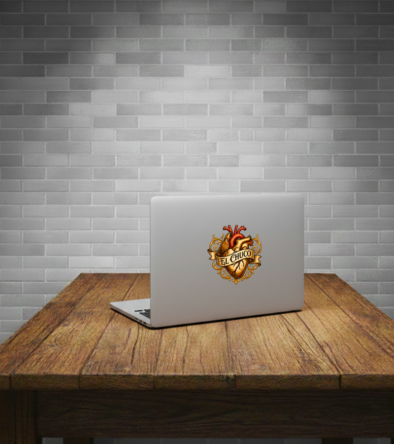 A silver laptop sits open on a rustic wooden table against a gray subway tile wall. The laptop lid features a prominent graphic decal of an anatomical heart with gold ornamental flourishes and the words 'BELIEVO' on a banner.