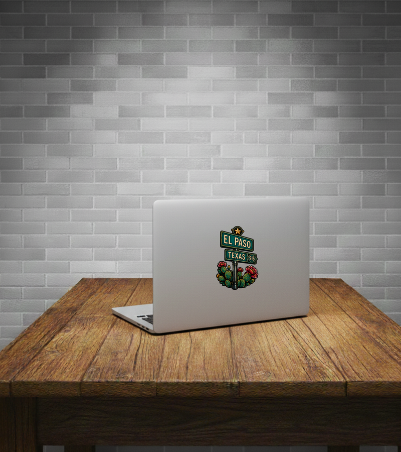 A silver laptop sits open on a rustic wooden table against a gray subway tile wall, displaying a colorful 'El Paso Texas' street sign sticker on the back.