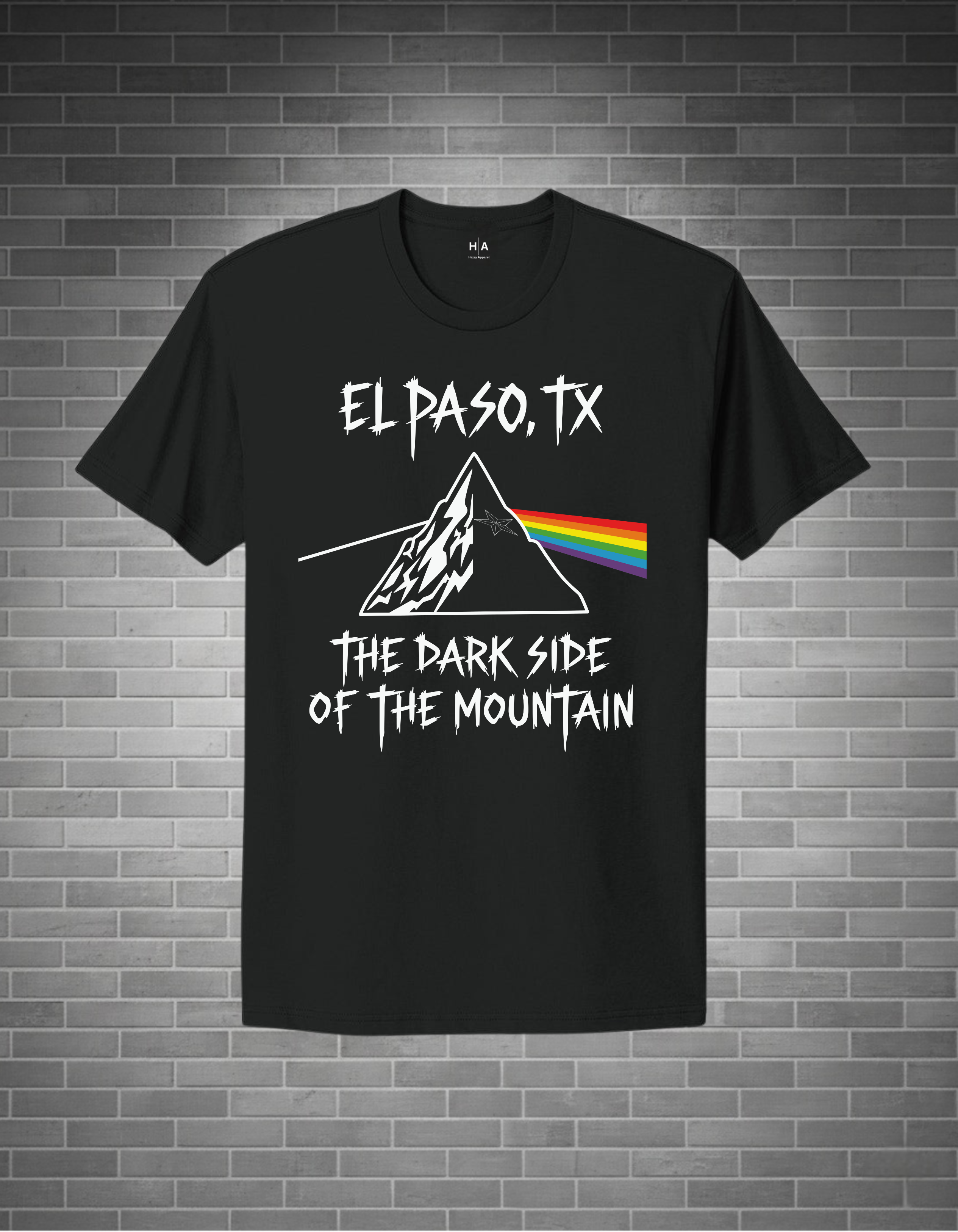 A black short-sleeve t-shirt with a white graphic print of a mountain and rainbow prism, accompanied by the text 'EL PASO, TX' and 'THE DARK SIDE OF THE MOUNTAIN', displayed against a gray brick wall background.