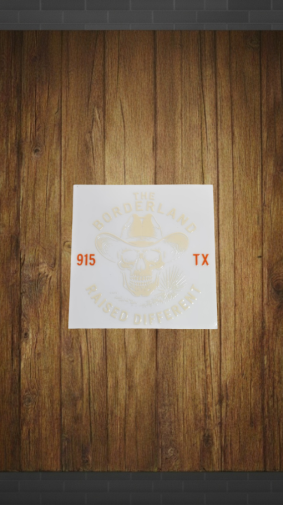 A square white sticker or decal with a faded cowboy skull graphic and orange text reading '915 TX', shown against a brown wood plank background.
