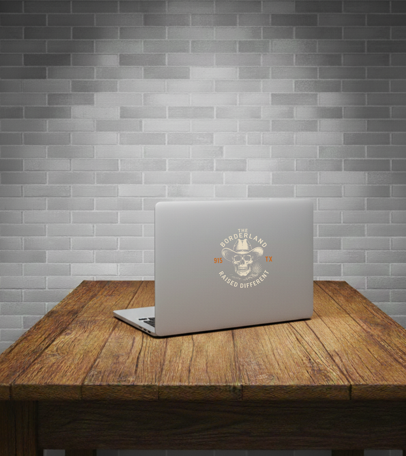 A silver laptop sits open on a rustic wooden table against a gray subway tile wall. The laptop lid has a circular graphic decal featuring a cowboy skull and the words 'WORLDLAND TRAIL RIDER HUNTER' in orange and gray.